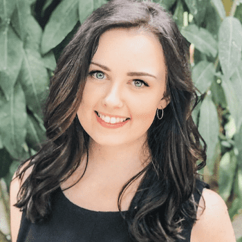 Emma Bostian, a young-looking White woman, smiling for the camera in front of green foliage.